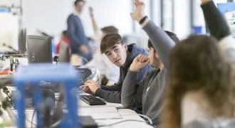 Des élèves de lycée en classe à table. Une rangée d'élèves qui lèvent la main pour prendre la parole et répondre à une question de leur professeur.
