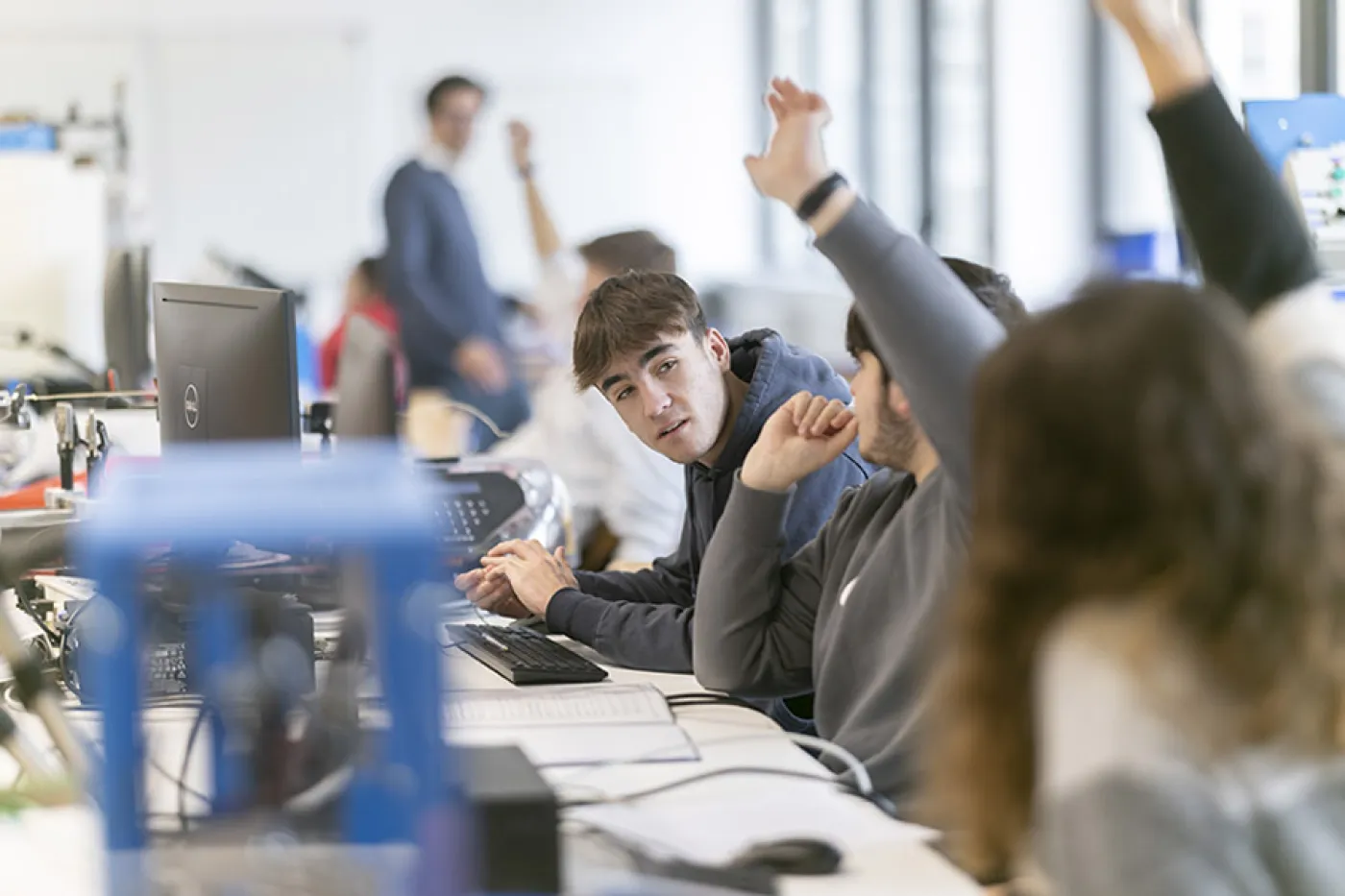 Des élèves de lycée en classe à table. Une rangée d'élèves qui lèvent la main pour prendre la parole et répondre à une question de leur professeur.