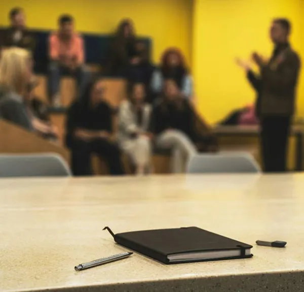 Un bureau dans une classe avec un carnet et, en arrière plan, un enseignant parle à un groupe d'étudiants